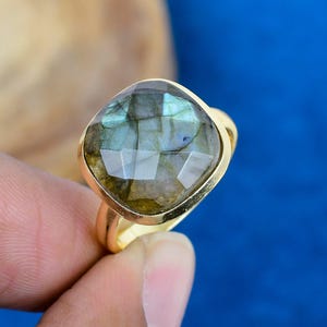 May include: A gold-toned ring featuring a large, faceted labradorite gemstone. The gemstone displays a mix of blue, green, and gray hues. The ring's setting is a simple, polished gold band, creating a classic and elegant look.
