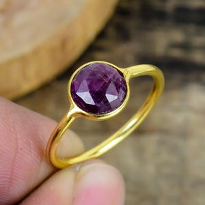 Op de afbeelding: Een gouden ring met een facetgeslepen, ronde, dieppaarse edelsteen. De ring heeft een eenvoudig, elegant ontwerp met een gladde, gepolijste gouden band. De edelsteen is gezet in een bezel setting.