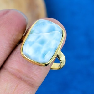 May include: A gold-toned ring featuring a rectangular, light blue gemstone with white marbling. The ring's band and setting are a bright gold color, creating a striking contrast with the stone's pale blue hue. The ring is held up by a finger.