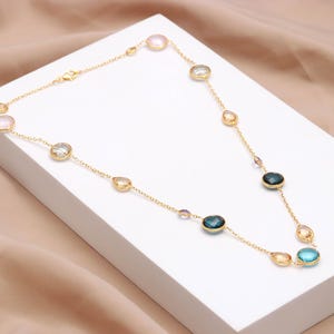 May include: A gold necklace with a delicate chain, featuring a series of colourful gemstones. The necklace is adorned with oval and round-cut stones in shades of pink, blue, and yellow. The necklace is displayed on a white surface.