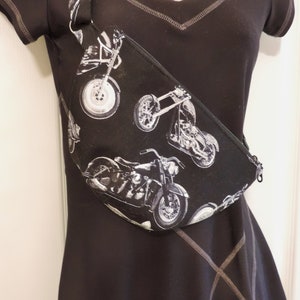 May include: A black fabric fanny pack with a white zipper and a motorcycle print. The fanny pack is worn diagonally across the body.