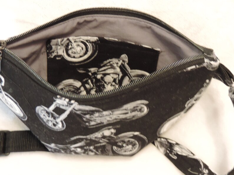 May include: Black zippered pouch with a gray lining and a black strap. The pouch is made of fabric with a repeating pattern of white motorcycles.