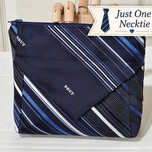 May include: A navy blue zippered pouch crafted from necktie fabric featuring diagonal white and light blue stripes. The pouch has a triangular flap with the word BRUT printed in white. A tag in the corner reads Just One Necktie with a tie icon.