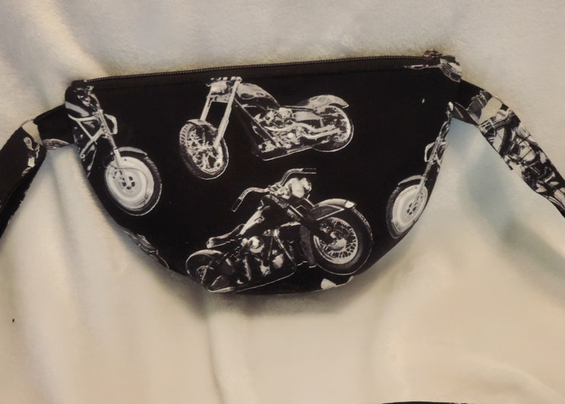 May include: Black fabric fanny pack with a zipper closure and a white print of motorcycles. The fanny pack has a long strap.