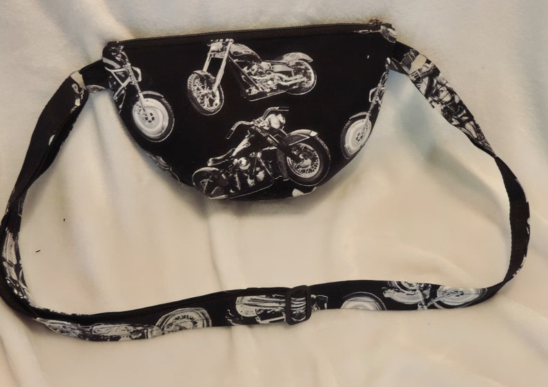 May include: Black fanny pack with a white adjustable strap. The fanny pack has a zipper closure and is decorated with a repeating pattern of white motorcycles.