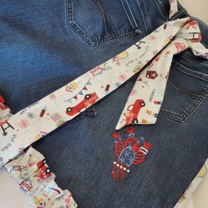 May include: A white fabric apron with a red, white, and blue Fourth of July theme. The apron has a tie-on waist and a ruffled bottom. The fabric features red trucks, fireworks, and the text "Land of the Free" and "Fourth of July".