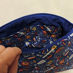 Puede incluir: Una bolsa azul con cremallera con un tirador de cremallera blanco. La bolsa est&aacute; forrada con tela azul y tiene un patr&oacute;n de herramientas y herrajes en el exterior.