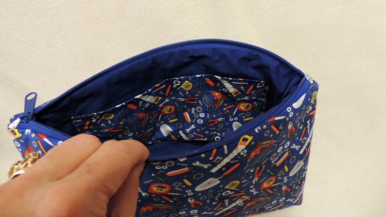 Puede incluir: Una bolsa azul con cremallera con un tirador de cremallera blanco. La bolsa est&aacute; forrada con tela azul y tiene un patr&oacute;n de herramientas y herrajes en el exterior.