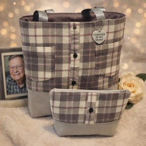 May include: A tote bag and matching zippered pouch made from a grey, white, and purple plaid fabric. The tote features two grey straps, two front pockets, and a central button placket. A heart-shaped charm hangs from a strap with the text FOREVER IN MY HEART.