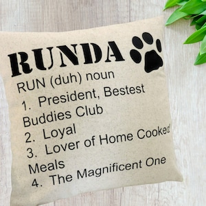 May include: A beige throw pillow with black text that reads "RUNDA RUN (duh) noun 1. President, Bestest Buddies Club 2. Loyal 3. Lover of Home Cooked Meals 4. The Magnificent One" and a black paw print.