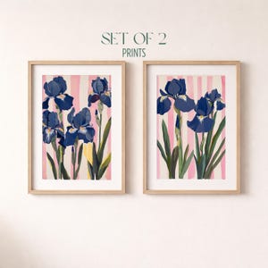 May include: Set of two framed art prints featuring watercolor illustrations of blue irises. The irises are set against a background of vertical pink and white stripes. The prints are in light brown wooden frames.