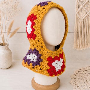 May include: A crochet hood featuring a mustard yellow base with a granny square pattern. The design incorporates floral-inspired motifs in vibrant red, white, and deep purple yarn, creating a textured, multi-colored look that covers the head and neck.