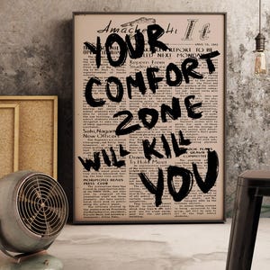 May include: A framed poster with the text "YOUR COMFORT ZONE WILL KILL YOU" in bold black letters. The poster is set against a vintage newspaper background. An old-fashioned fan and a small framed picture are in the foreground.