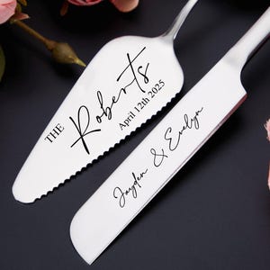 Könnte beinhalten: Ein silbernes Tortenheber- und Messerset mit schwarzer Schrift. Auf dem Servierer steht "The Roberts" und "April 12th 2025". Auf dem Messer steht "Jayden & Evelyn". Das Set liegt auf einer dunklen Oberfläche mit rosa und roten Blumen.