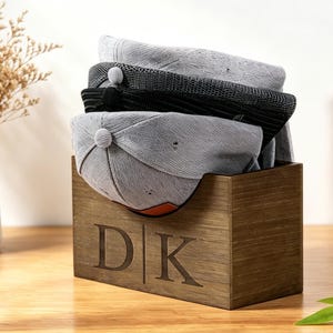 Puede incluir: Una caja de madera con las iniciales "DK" grabadas contiene varias gorras de b&eacute;isbol. Las gorras son grises y negras, con un bot&oacute;n en la parte superior. La caja es de madera oscura y est&aacute; sobre una superficie de madera.