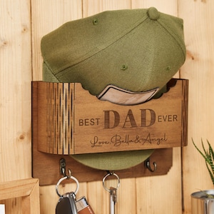 Op de afbeelding: Een houten hoeden- en sleutelhouder met de tekst "BEST DAD EVER" en de namen "Love, Bella & Angel" gegraveerd. Een olijfgroene baseballcap staat in de houder, met sleutels eronder. De houder is op een houten muur gemonteerd.