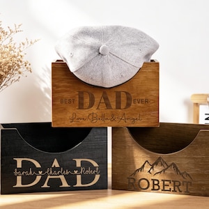 Puede incluir: Tres cajas de madera con texto grabado. La caja superior es de color marr&oacute;n claro y dice "BEST DAD EVER" con una gorra gris en su interior. La caja del medio es negra y dice "DAD". La caja inferior es marr&oacute;n y dice "ROBERT" con un gr&aacute;fico de monta&ntilde;a.