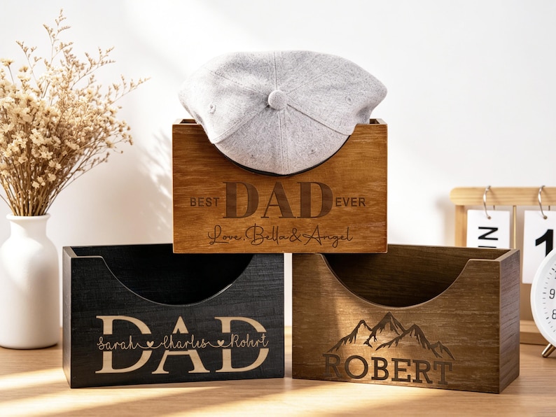 Puede incluir: Tres cajas de madera con texto grabado. La caja superior es de color marr&oacute;n claro y dice "BEST DAD EVER" con una gorra gris en su interior. La caja del medio es negra y dice "DAD". La caja inferior es marr&oacute;n y dice "ROBERT" con un gr&aacute;fico de monta&ntilde;a.