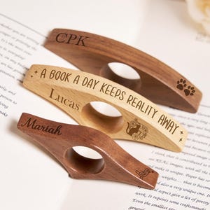 May include: Three wooden book page holders with engraved text and designs. One is light wood with "A BOOK A DAY KEEPS REALITY AWAY" and "Lucas". Another is dark wood with "Mariah". The third is dark wood with "CPK".