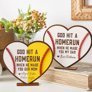 Könnte beinhalten: Zwei herzförmige Holzschilder mit Baseballnähten und dem Text "GOD HIT A HOMERUN". Eine Tafel hat ein gelbes Baseball-Design, die andere ein weißes. Ein Baseball und Blumen sind ebenfalls im Bild.