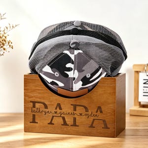 Puede incluir: Tres gorras de b&eacute;isbol en una caja de madera con la palabra "PAPA" grabada en la parte delantera. Las gorras son grises y negras, con un estampado de camuflaje en la parte delantera de una. Un peque&ntilde;o jarr&oacute;n con flores secas y un calendario de madera est&aacute;n en el fondo.