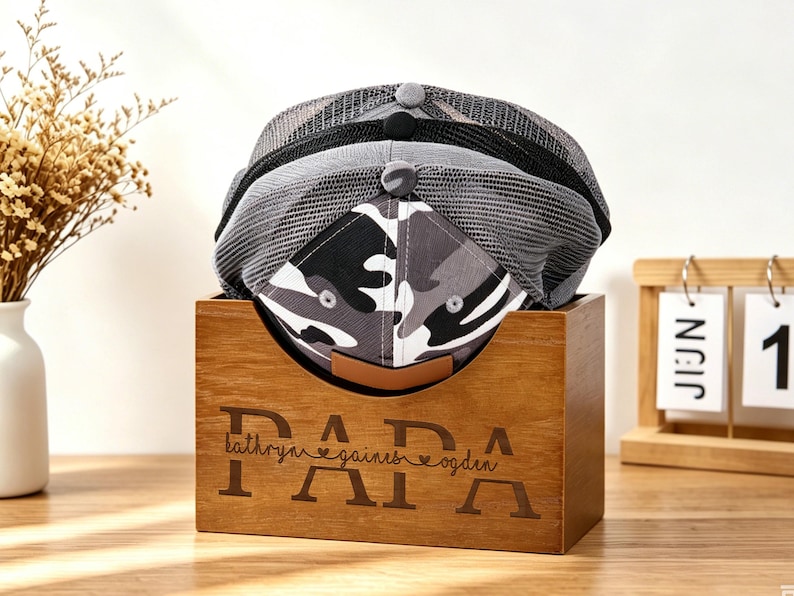 Puede incluir: Tres gorras de b&eacute;isbol en una caja de madera con la palabra "PAPA" grabada en la parte delantera. Las gorras son grises y negras, con un estampado de camuflaje en la parte delantera de una. Un peque&ntilde;o jarr&oacute;n con flores secas y un calendario de madera est&aacute;n en el fondo.