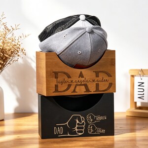 Puede incluir: Dos gorras de b&eacute;isbol descansan en una caja de almacenamiento de madera. La caja superior est&aacute; grabada con la palabra "DAD" y nombres. La caja inferior es negra con un gr&aacute;fico de pu&ntilde;o y nombres. Un jarr&oacute;n de flores secas y un calendario est&aacute;n en el fondo.