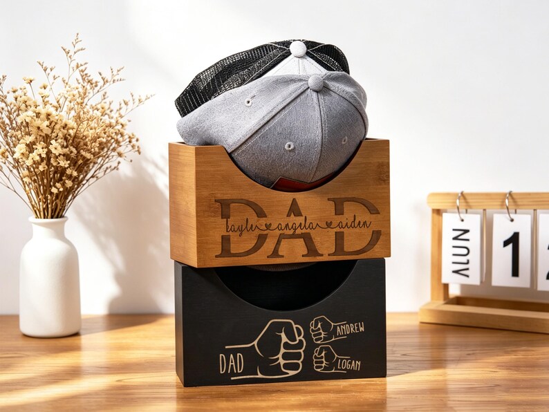 Puede incluir: Dos gorras de b&eacute;isbol descansan en una caja de almacenamiento de madera. La caja superior est&aacute; grabada con la palabra "DAD" y nombres. La caja inferior es negra con un gr&aacute;fico de pu&ntilde;o y nombres. Un jarr&oacute;n de flores secas y un calendario est&aacute;n en el fondo.