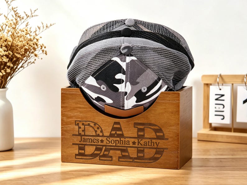 Puede incluir: Una caja de madera con la palabra "DAD" grabada y los nombres James, Sophia y Kathy, que contiene tres gorras de b&eacute;isbol. Las gorras tienen patrones de malla gris y negra y camuflaje. La caja es de madera.