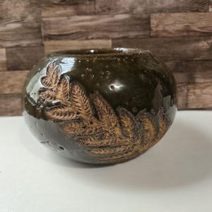 May include: A round, dark olive green ceramic bowl with a glossy finish. The exterior features a textured, light brown fern leaf imprint that wraps around the side. The bowl has a wide, open top and a smooth, rounded base with visible surface details.