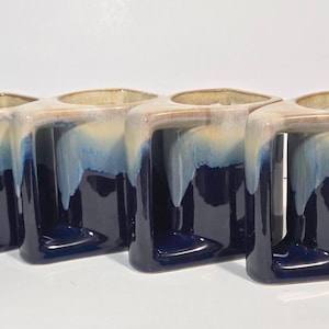 May include: A set of four ceramic mugs featuring a distinct geometric shape with a flat top and a wide, angular handle. The mugs have a two-tone glaze, transitioning from a deep, glossy navy blue at the base to a creamy, light beige at the rim.