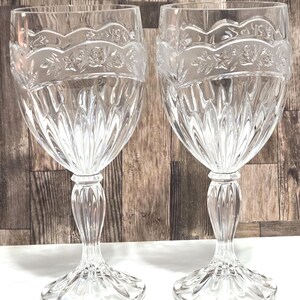 May include: Two clear crystal wine glasses with ornate floral and leaf designs etched around the rim. The glasses have a tulip-shaped bowl and a detailed stem and base. The background is a wooden surface.