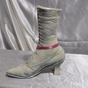 May include: A vintage-style, pale green boot with a pointed toe and a small heel. The boot features a lace-up design and a decorative pink ribbon around the ankle. The boot is made of a soft material and has a worn appearance.