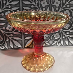 May include: A pedestal bowl crafted from glass featuring a vibrant gradient of amber and deep red. The bowl has a wide, shallow basin with a diamond-patterned exterior and a beaded rim, supported by a thick, faceted stem and a circular base.