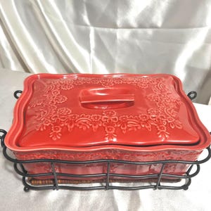 May include: A red ceramic casserole dish with a lid, featuring a floral embossed design. The dish is set in a black metal wire frame with handles. The dish is approximately 12 inches long and 8 inches wide.