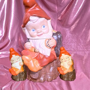May include: A ceramic gnome cookie jar with two smaller gnome figurines. The cookie jar gnome wears a red hat and orange outfit, holding a cookie. The smaller gnomes have orange hats and outfits. All have white beards and blue eyes.