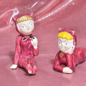 May include: Two ceramic figurines of children in red hooded outfits with yellow hair and white faces. One is seated, the other is lying down. The figurines have blue eyes and are on a pink background.