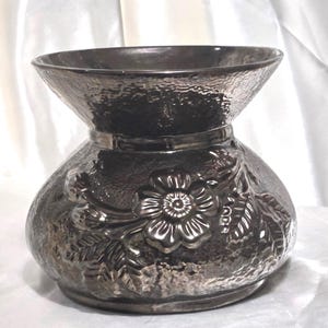 May include: A silver-toned vase with a textured surface and a flared top. The vase features a floral design in relief around the base. The vase has a band around the middle and a wide opening.