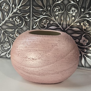 May include: A round, blush-colored vase with a textured surface. The vase has a wide opening and sits in front of a decorative silver panel with floral designs. The vase's exterior has a speckled appearance.