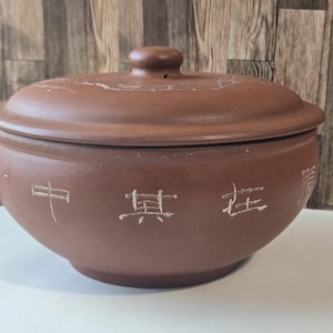 May include: A brown ceramic cooking pot with a lid. The pot has a rounded shape with a handle on one side and Chinese characters etched into the side. The lid has a small knob on top. The pot is designed for cooking.