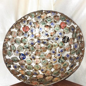 May include: A round, handcrafted bowl made from recycled soda can tabs. The bowl features a metal frame and is filled with colorful, vintage soda can tabs, creating a unique, textured surface. The tabs display various brand logos.