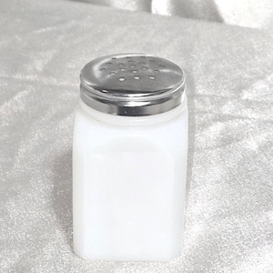 May include: A white, rectangular salt shaker with a silver metal lid. The lid has small holes for dispensing salt. The shaker is made of opaque white glass or ceramic.