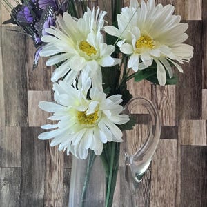 May include: A clear glass pitcher holds a bouquet of white daisies with yellow centers and green stems. Purple butterfly decorations and tall green grass accents the flowers. The pitcher sits against a wooden background.