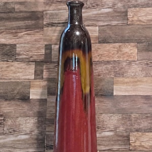 May include: A tall, slender ceramic vase with a glossy finish. The base is a deep, solid red that transitions upward into a dark brown neck. A distinct drip-glaze effect in shades of amber and gold creates a vertical pattern where the colors meet.