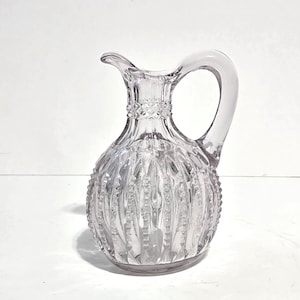 May include: A clear glass pitcher featuring a rounded body with vertical ribbed patterns and a textured diamond band around the neck. The vessel includes a curved handle and a small pouring spout, showcasing a transparent and reflective surface.