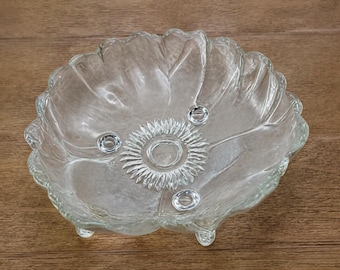 Puede incluir: Un cuenco de cristal transparente con borde festoneado y un patrón moldeado en forma de hoja. La base interior muestra un diseño central de rayos de sol rodeado por tres pequeñas hendiduras circulares. El cuenco se apoya sobre tres patas cortas.