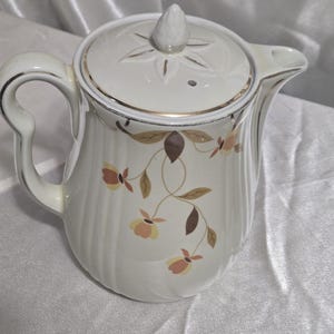 May include: Cream-colored ceramic teapot with a lid and a curved handle. The teapot features a floral design in shades of brown and peach, with gold trim around the rim and lid. The lid has a star-shaped design.
