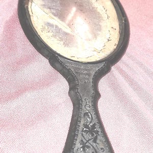 May include: An antique hand mirror with a dark metal frame and handle. The oval mirror has a cracked surface. The handle is decorated with floral designs and the text "& MARON 19 1872".