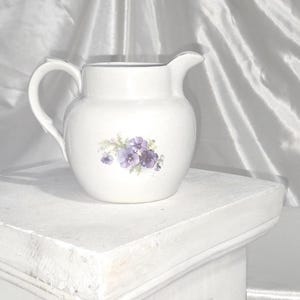 May include: White ceramic pitcher with a handle and spout, decorated with a cluster of purple flowers and green leaves. The pitcher is sitting on a white surface against a white background.
