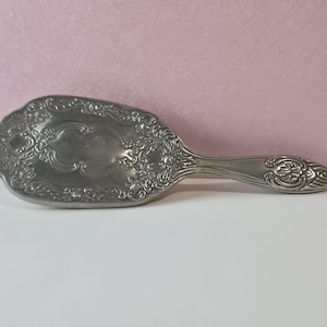 May include: An antique silver-toned hairbrush with an ornate, floral design. The brush has a long handle and a wide, oval head. The handle and head are decorated with detailed scrollwork and floral patterns.